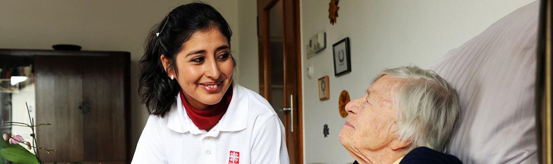 Caritas-Mitarbeiterin an Bett einer pflegebedürftigen Frau