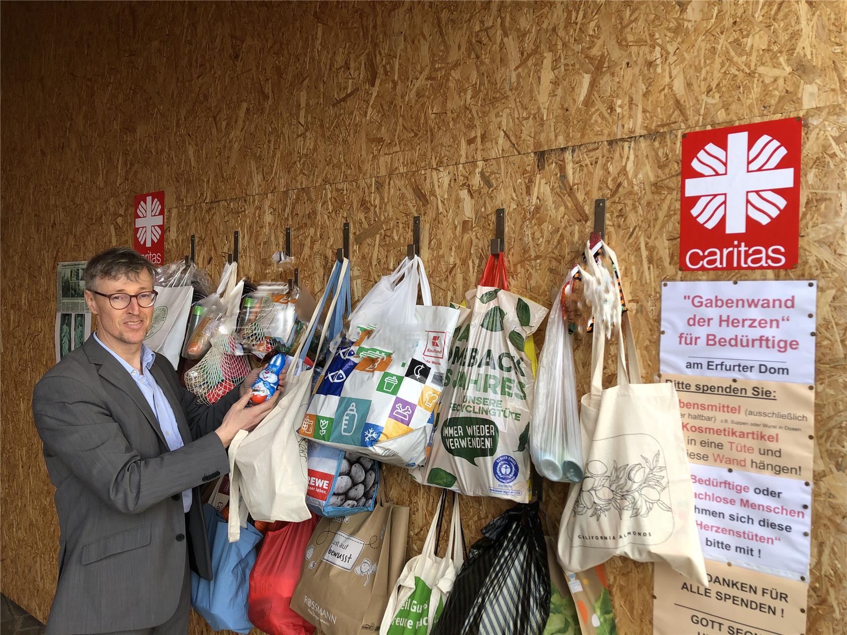 Caritasdirektor Wolfgang Langer steht an der „Gabenwand der Herzen“, die mit zahlreichen bunten Tüten voller Spenden bestückt ist