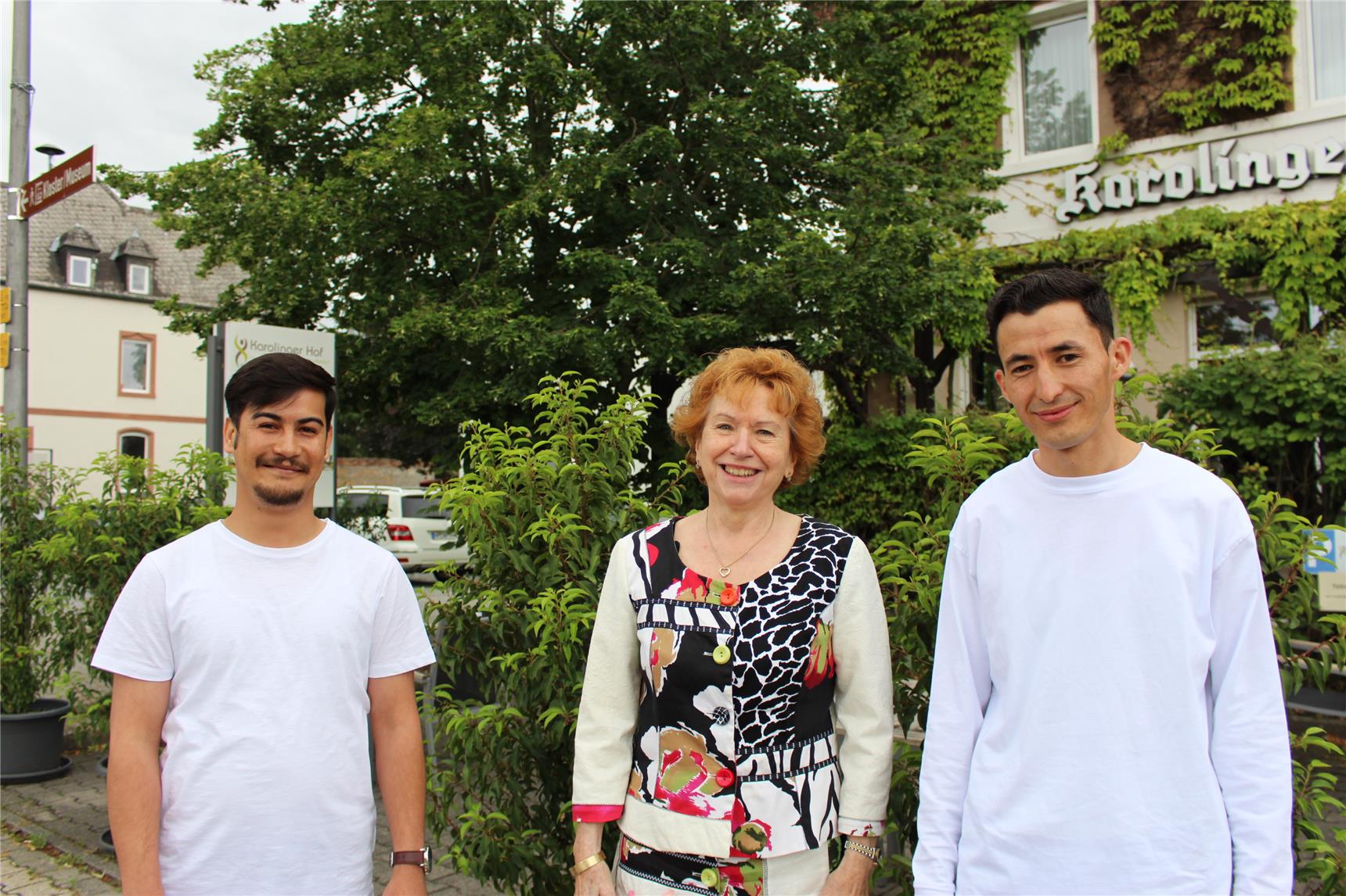 Eine Same steht vor einem Gebüsch und blickt in die Kamera. Links und rechts neben ihr stehen zwei junge Männer. 