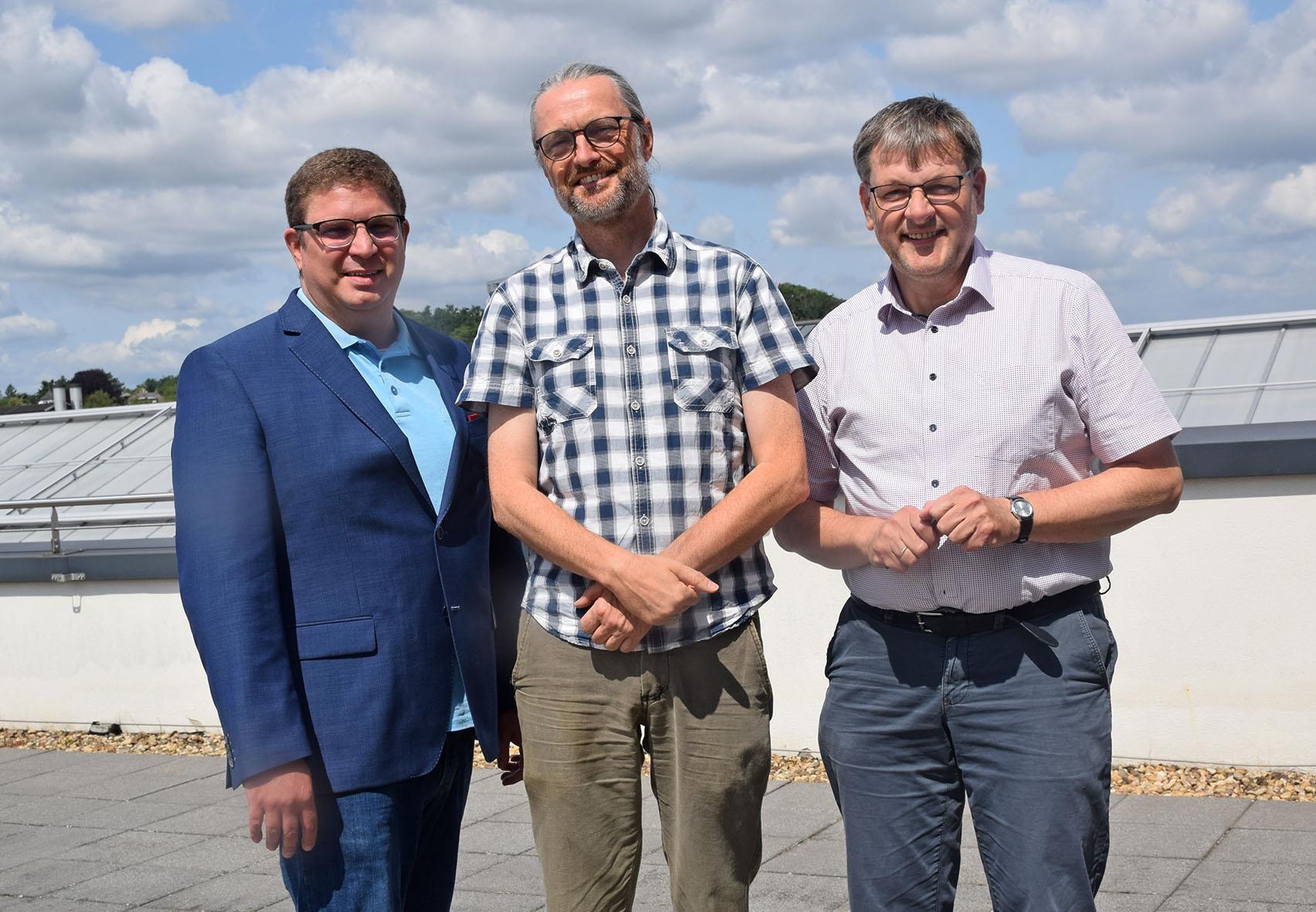 Drei Männer stehen auf einer Terrasse udn lächeln in die Kamera.