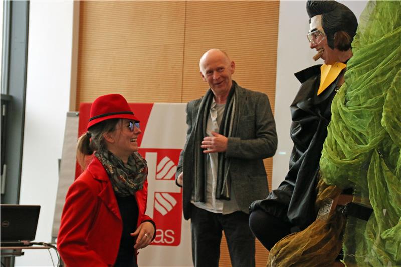 Zwei verkleidete Frauen spielen Theater. Im Hintergrund beobachtet ein Mann das Spiel.