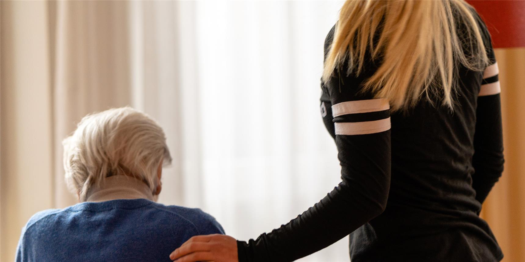 Zwei Frauen sind von hinten zu sehen, die alte Dame mit grauem Haar sitzt, eine junge Frau steht neben ihr und legt die Hand auf ihre Schulter.