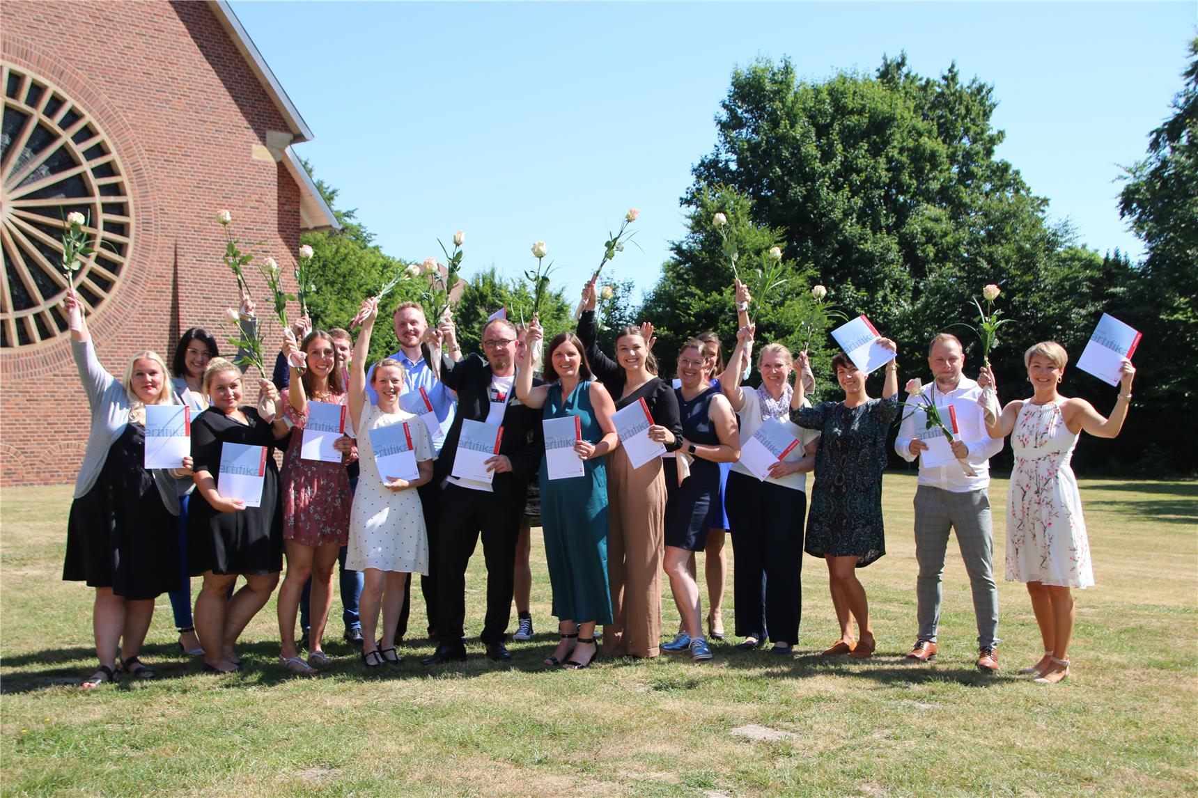 Im Freien auf einer grünen Wiese stehen 17 Kursteilnehmer*innen mit Zertifikatmappen und Rosen, die sie in die Luft strecken