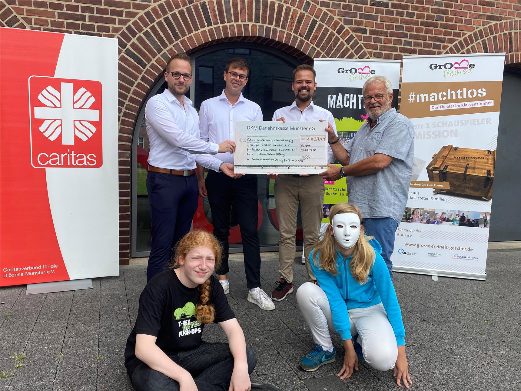 Vier Männer stehen beisammen und halten einen großen Scheck in der Hand, vor ihnen sitzen zwei Kinder auf dem Boden. Eines trägt eine Theatermaske.