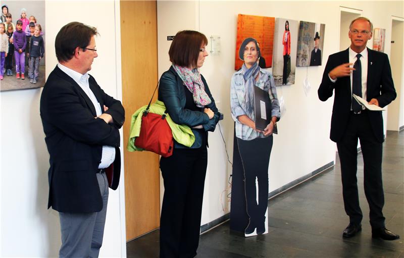 Drei Menschen stehen in einem Raum. Die Person ganz rechts erzählt etwas und gestikuliert dabei. Die zwei anderen hören zu.