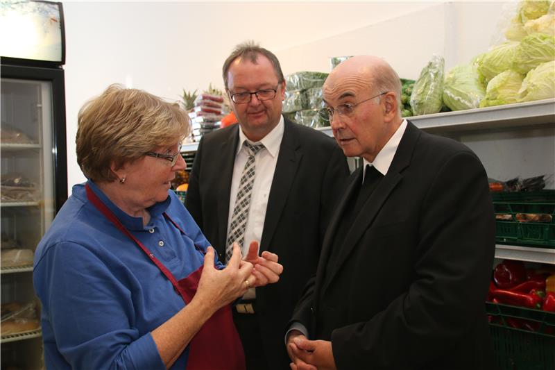 Eine Frau und zwei Männer reden miteinander. Rechts von ihnen steht ein Regal mit Gemüse und links ein Kühlschrank mit Glastür.