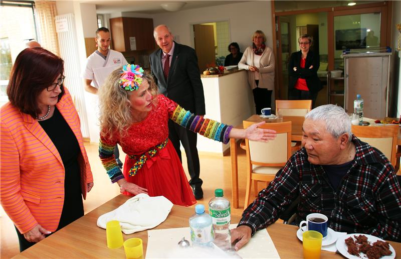 Das Foto zeigt eine Clownin mit einem einem Altenheimbewohner und drumherum Zuschauer.