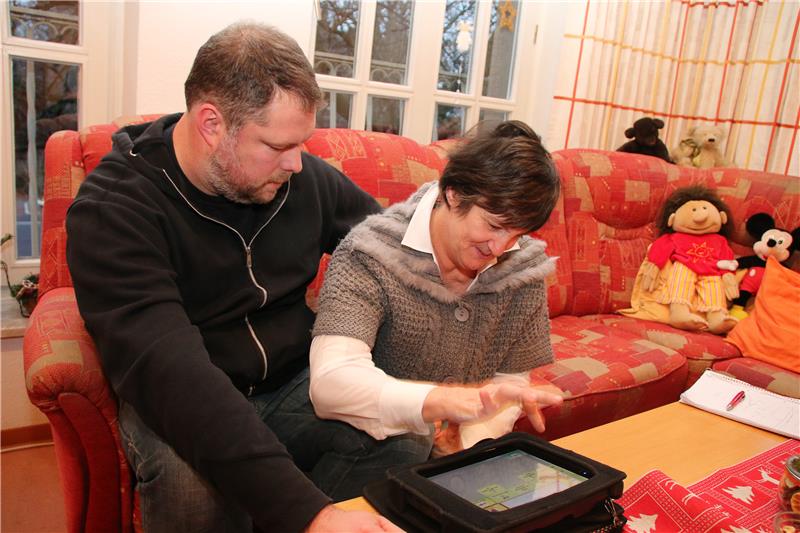 Das Foto zeigt einen Mann, der neben einer behinderten Frau auf einem Sofa sitzend ihr hilft, einen Tablet-Computer zu bedienen.