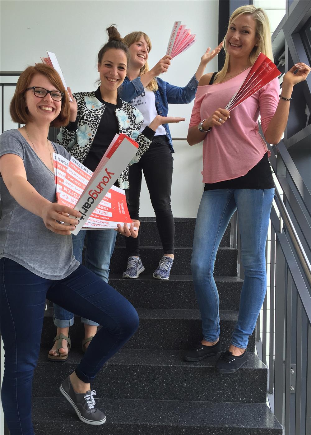 Vier junge Damen stehen versetzt auf einer Treppe und halten fächerförmige Pappen in die Kamera.