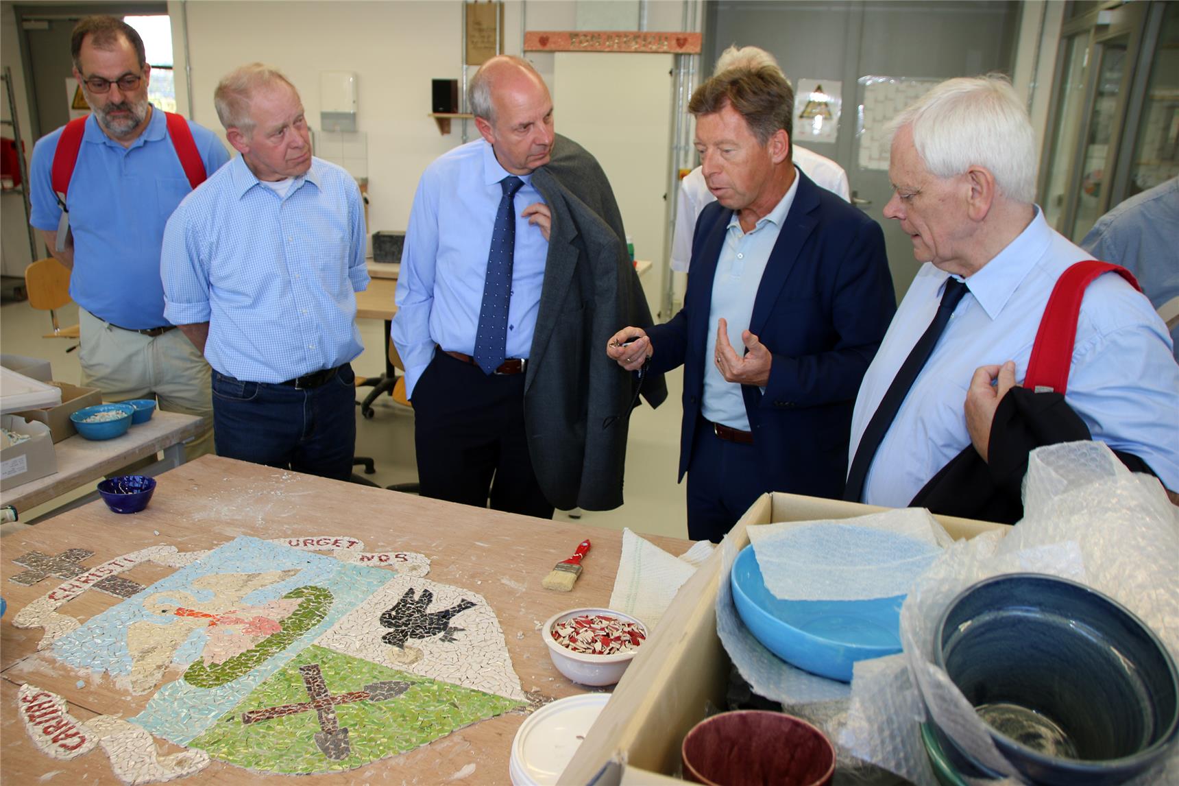 Das Foto zeigt eine kleine Gruppe von Menschen vor einem Werkstatttisch, auf dem ein Mosaik entsteht.