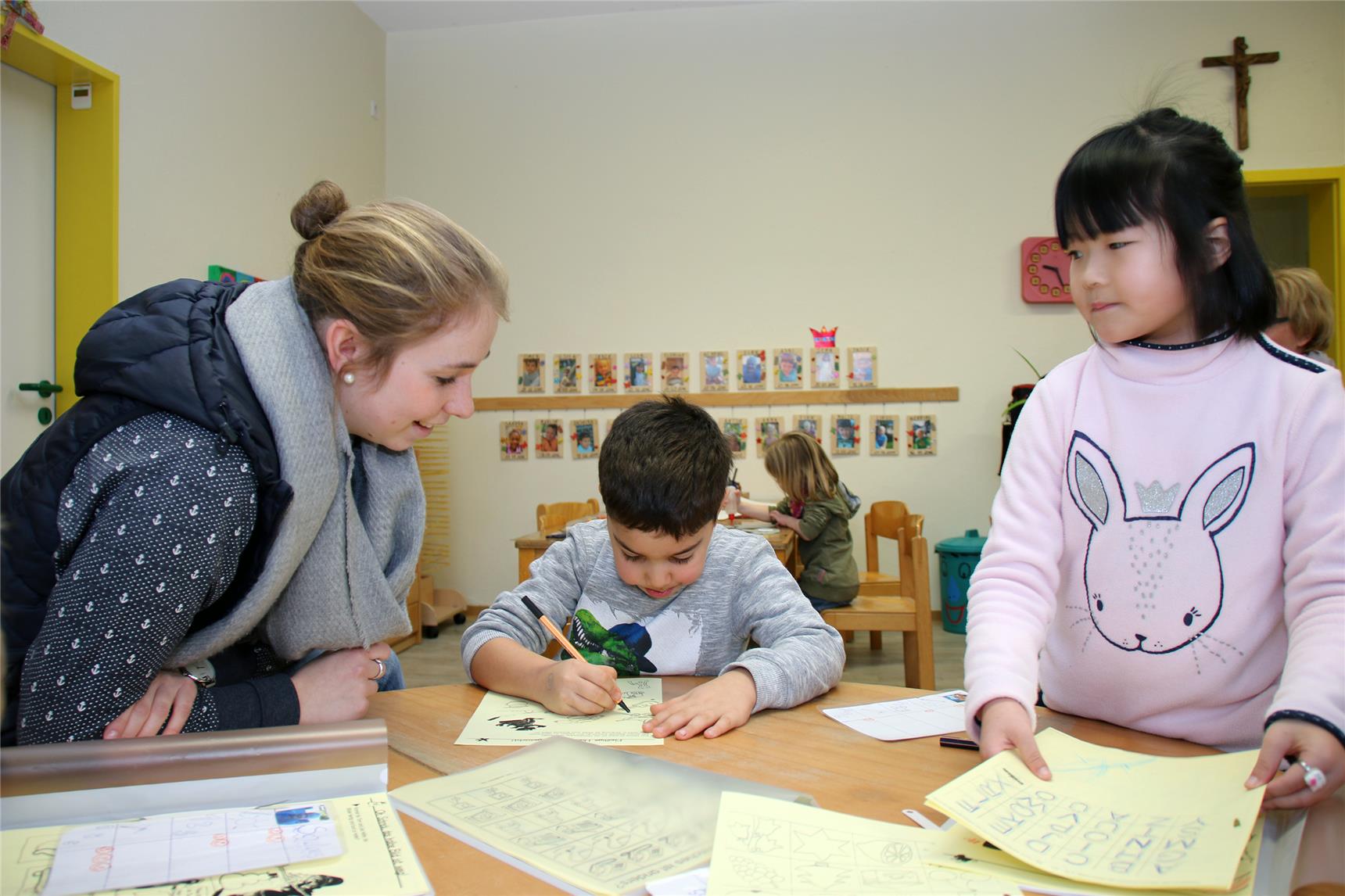 Am Tisch malen zwei Kinder, eine Erzieherin sitzt dabei