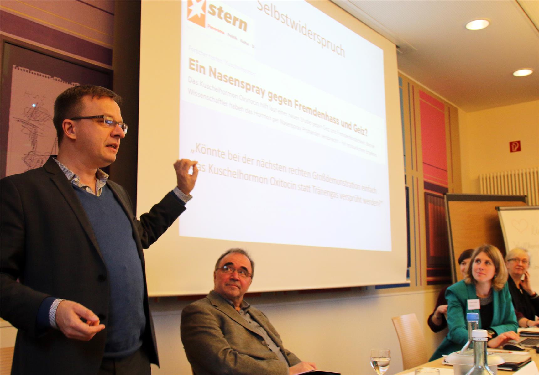 Auf dem Foto sitzen einige Menschen an Tischen, ein Mann steht vor der Leinwand