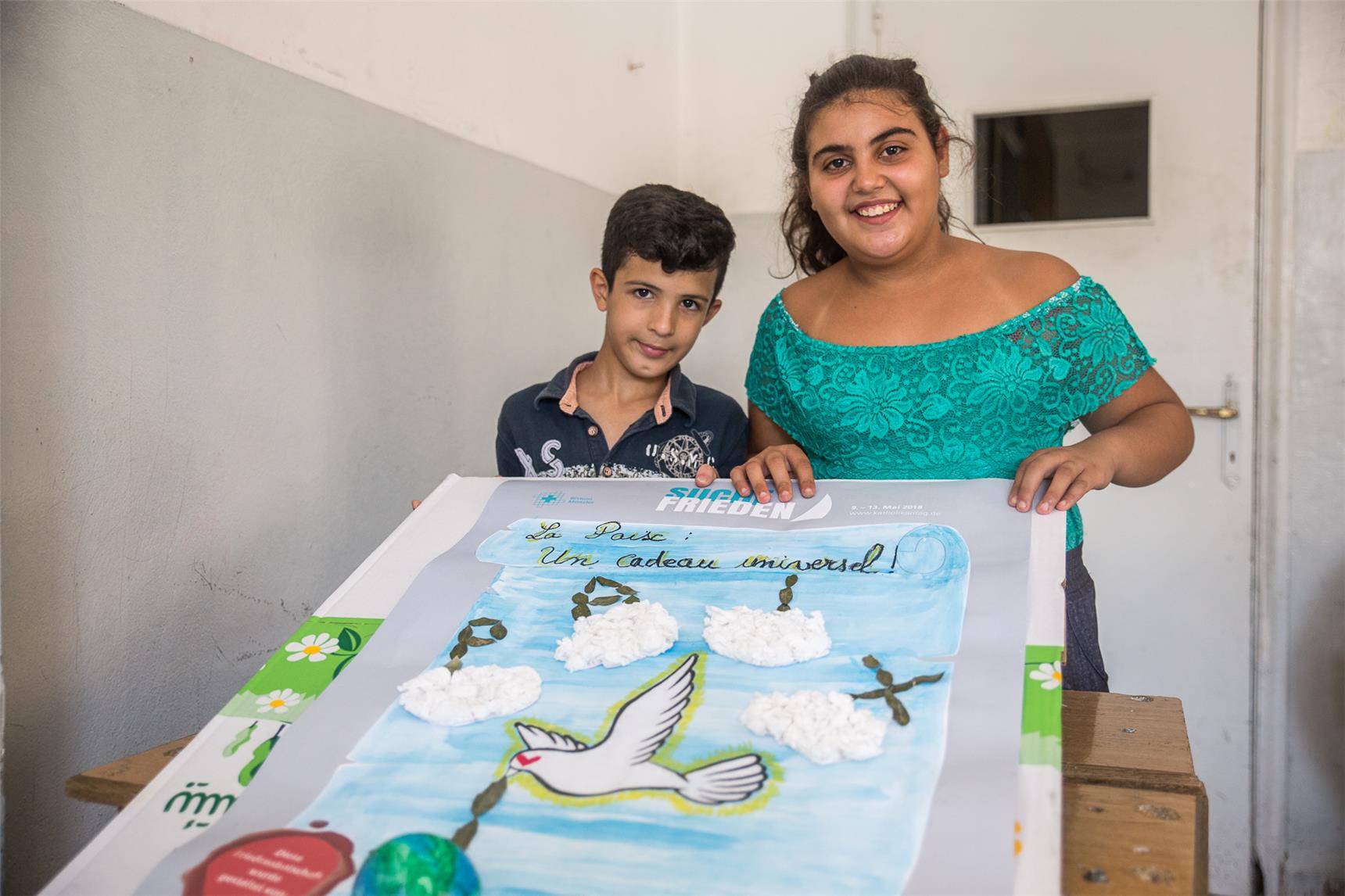 Zwei syrische Flüchtlingskinder präsentieren den von ihnen gestalteten Friedensvertrag mit Wolken, Olivenzweigen und einer kleinen Weltkugel