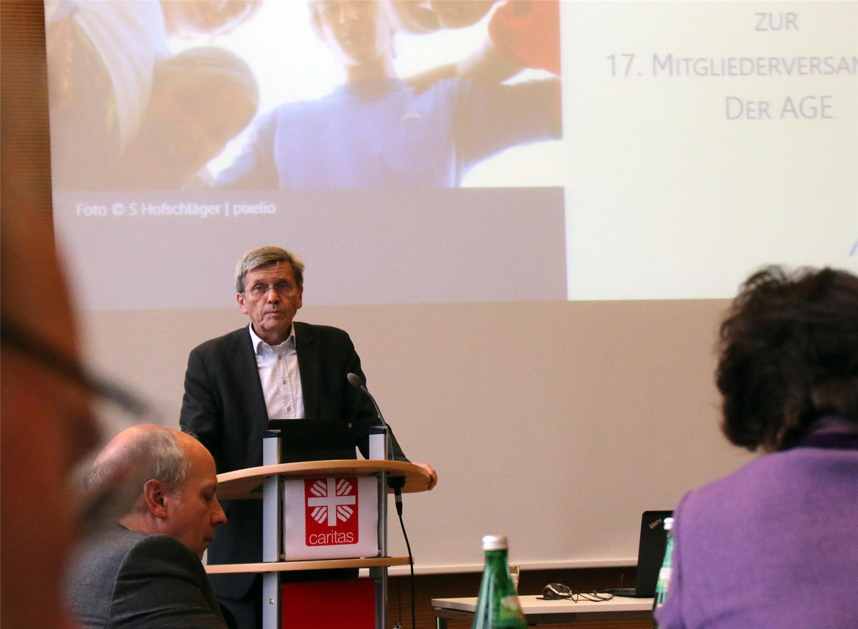 Das Foto zeigt einen Redner am Pult durch einige Zuhörer hindurch fotografiert.