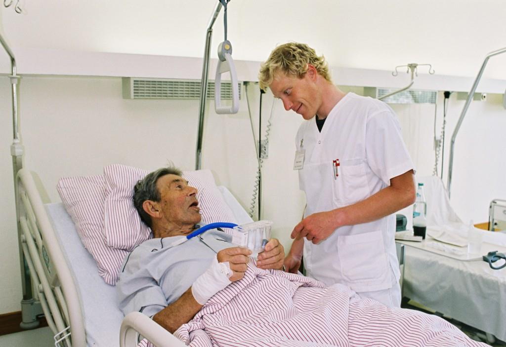 Das Foto zeigt einen Krankenpfleger, der mit einem Patienten im Krankenbett spricht.