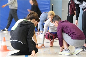 039-2019_Bewegung_OGS.pg Vier Personen hocken in einer Turnhalle und schauen sich etwas auf dem Boden an.