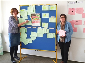 031-2020 Koordinierungsstelle Zwei Frauen stehen in einem Raum, in ihrer Mitte eine Magnettafel. An diese sind grüne Zettel geheftet mit Corona-Hilfsangeboten.