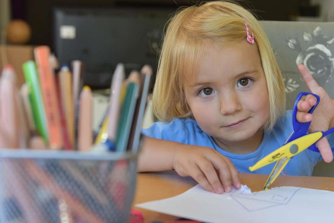 Ein Kind mit Bastelschere sitzt an einem Tisch und schaut in die Kamera