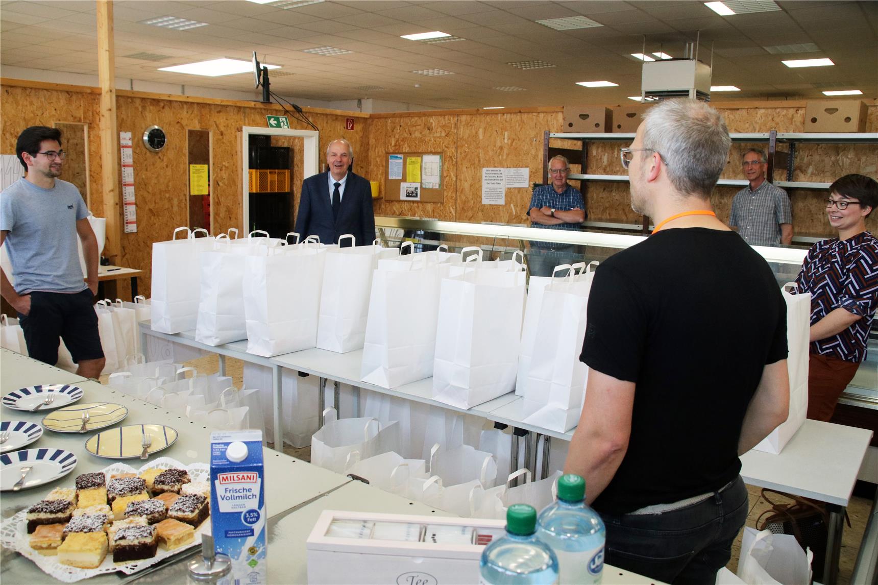 Das Foto zeigt eine weit auseinanderstehende Gruppe von Menschen in einem Raum, in dem viele weiße Tüten aufgestellt sind.