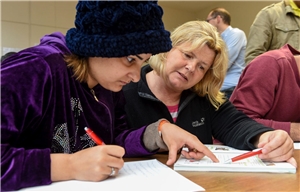 058-2020 Arbeitslosenreport Das Foto zeigt eine Frau und eine Mann, die unterstültzt von einer Frau in der Mitte konzentriert in Hefte schreiben.