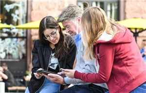073-2020 Förderung Fernsehlotterie Zwei Frauen und ein Mann sitzen in einem Außencafé und schauen auf ein tablet.