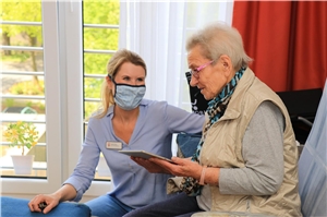 083-2020_Pflegelohn Eine Frau schaut eine ältere Dame an, die in einem Sessel sitzt und ein Tablet in ihren Händen hält.