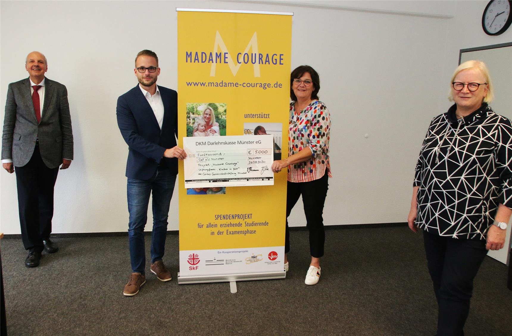 Vier Personen stehen in einem Raum. Zwei von ihnen halten einen Scheck der DKM Darlehenskasse Münster eG in den Händen.