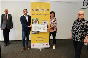 088-2020_Spende_Madame-Courage Vier Personen stehen in einem Raum. Zwei von ihnen halten einen Scheck der DKM Darlehenskasse Münster eG in den Händen.