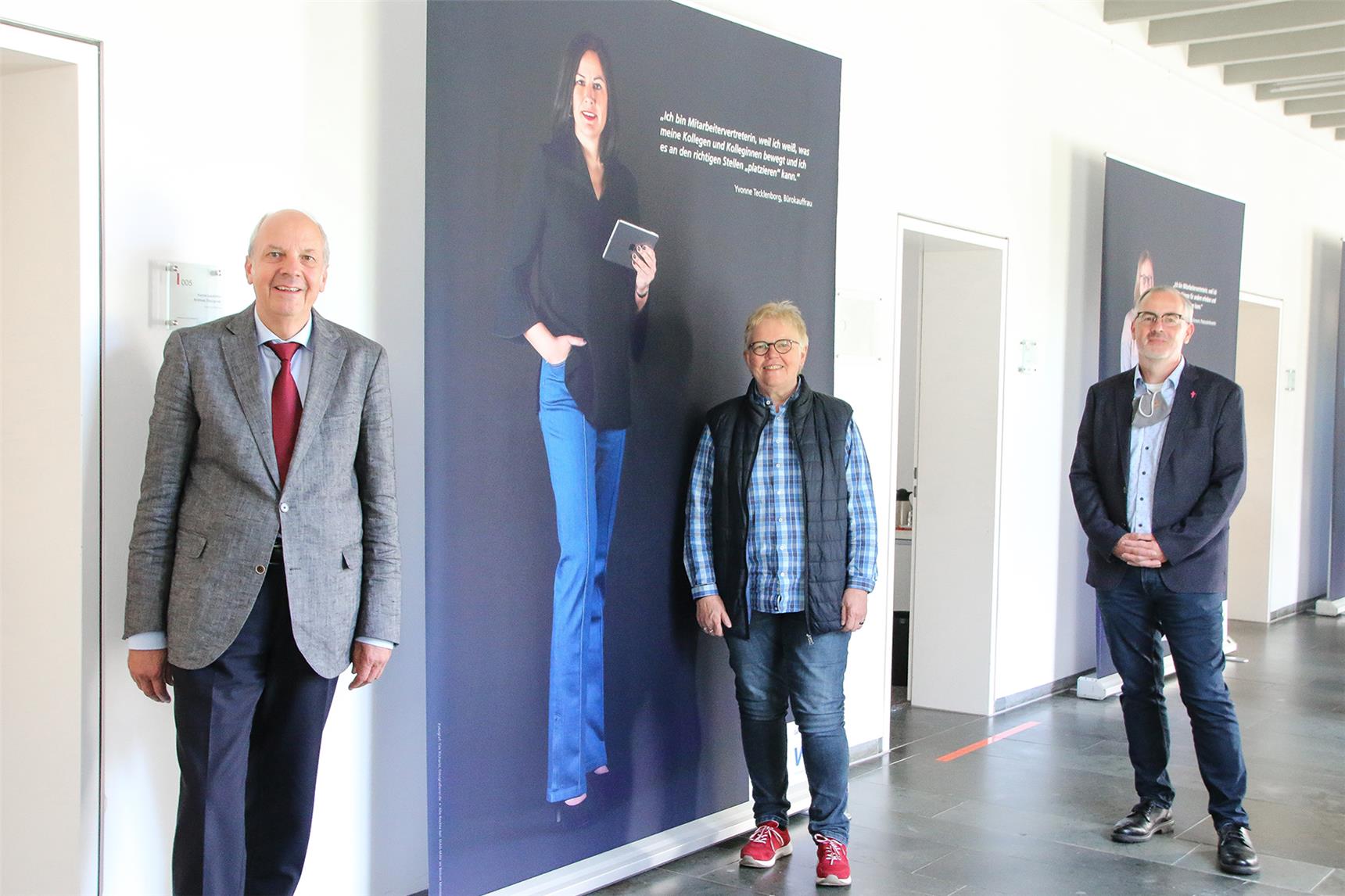 Drei Personen stehen mit Abstand in einem Flur, hinter ihnen ein zirka zwei Meter hohes Transparent, auf dem eine Frau und Text abgebildet sind