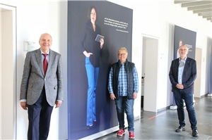 090-2020 Ausstellungseröffnung MAV Drei Personen stehen mit Abstand in einem Flur, hinter ihnen ein zirka zwei Meter hohes Transparent, auf dem eine Frau und Text abgebildet sind