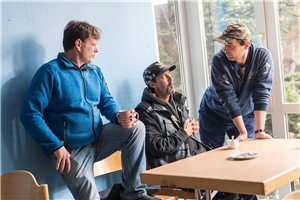 109-2020 Armut und Migration Das Foto zeigt drei Männer, die sich an einem Tisch stehend und sitzend unterhalten.