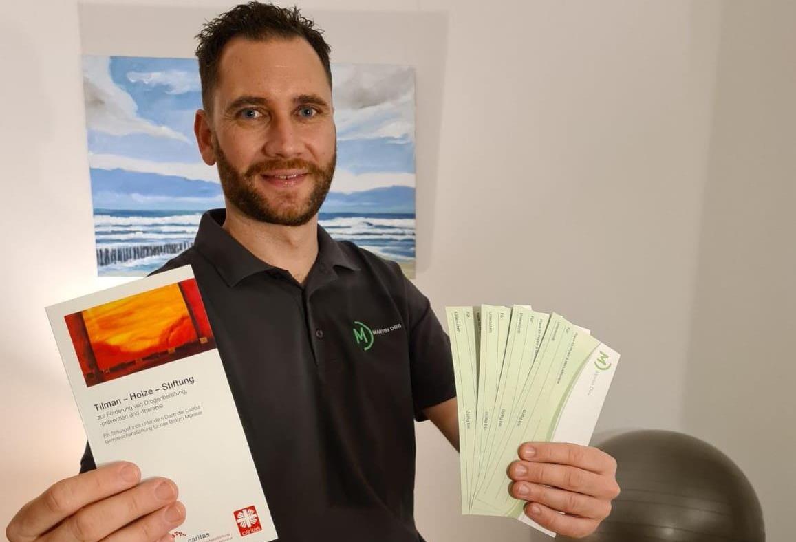 Ein Mann sitzt auf einer Liege und lächelt in die Kamera. In einer Hand hält er einen Flyer der Tilman-Holze-Stiftung, in der anderen die aufgefächert einige Gutscheine.