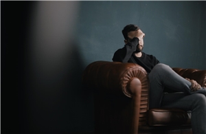 004-2022 Täterprojekt Das Foto zeigt einen verzweifelten Mann auf einer Couch in einem dunklen Raum.