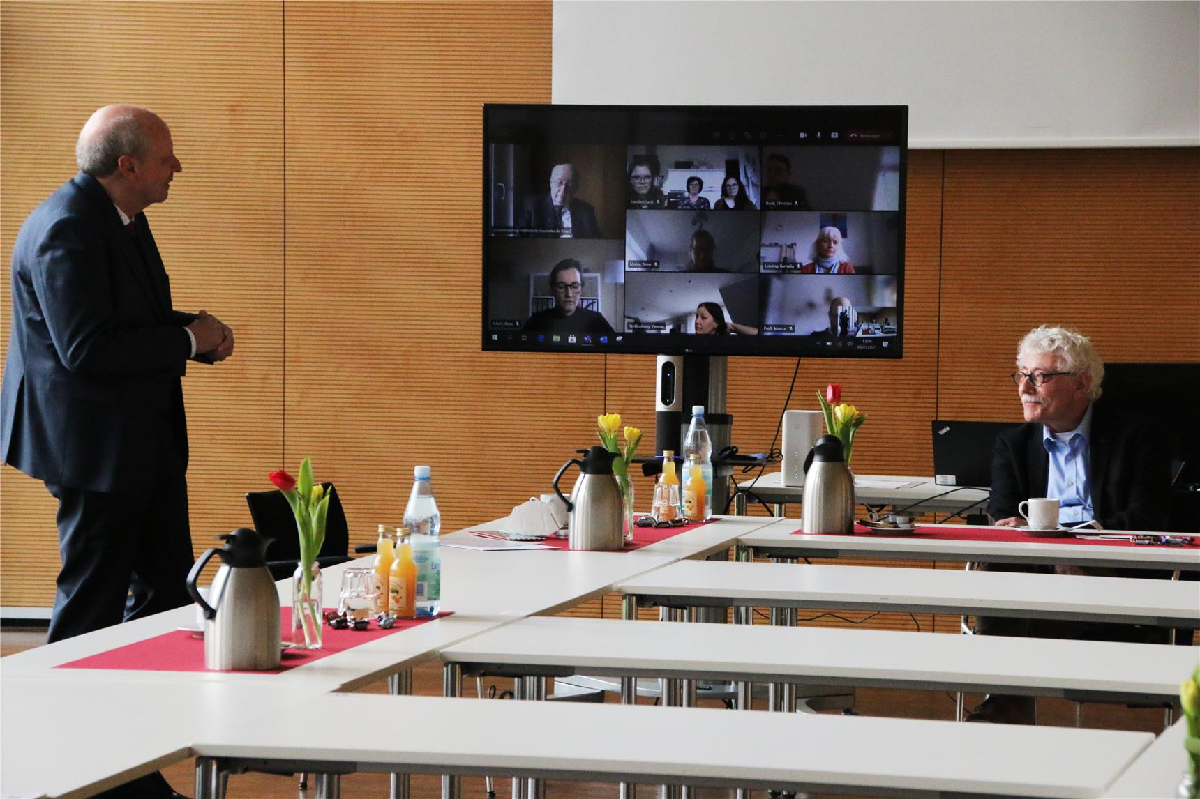 Das Foto zeigt zwei Männer in einem Großen Raum und dazwischen ein Bildschirm, auf dem eine Videokonferenz zu sehen ist.