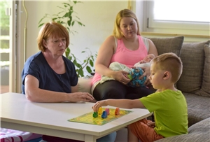 031-2021 Frühförderung Das Foto zeigt zwei Frauen an einem Wohnzimmertisch, die mit einem Jungen ein Brettspiel spielen.