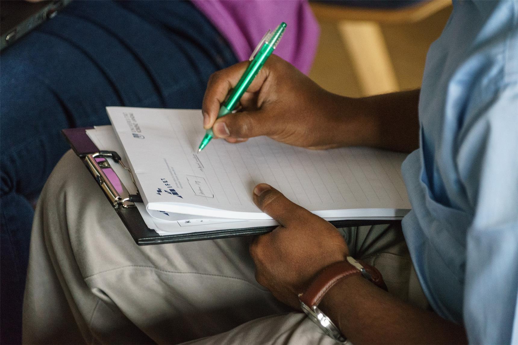 Jemand sitzt auf einem Stuhl, nur die überschlagenen Beine sind sichtbar, darauf liegt ein Klemmbrett mit Block, auf dem die Person schreibt