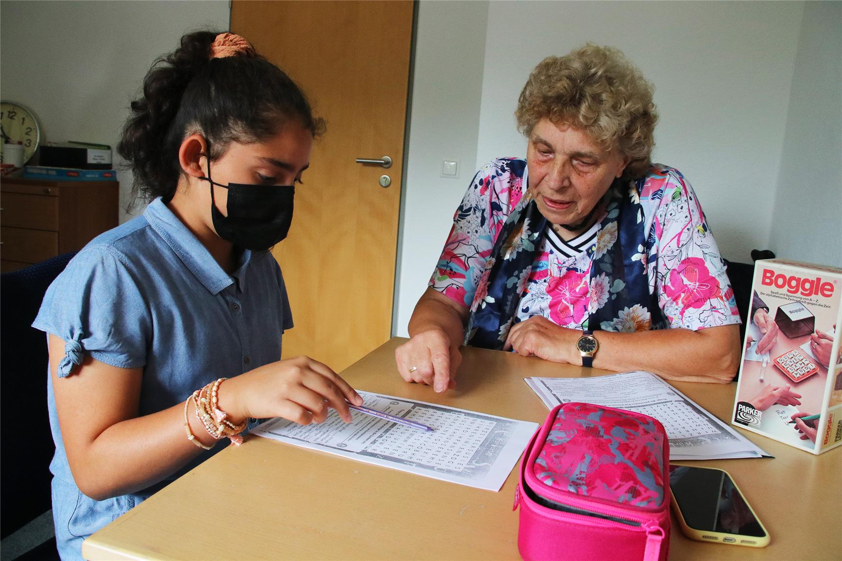 Das Foto zeigt eine ältere Frau, die einem jungen Mädchen bei Hausaufgaben hilft.