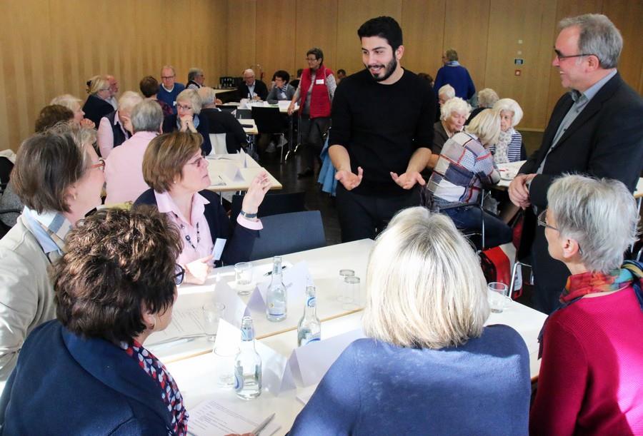 Zwei Männer stehen im Raum und diskutieren mit fünf sitzenden Personen an einem Tisch.