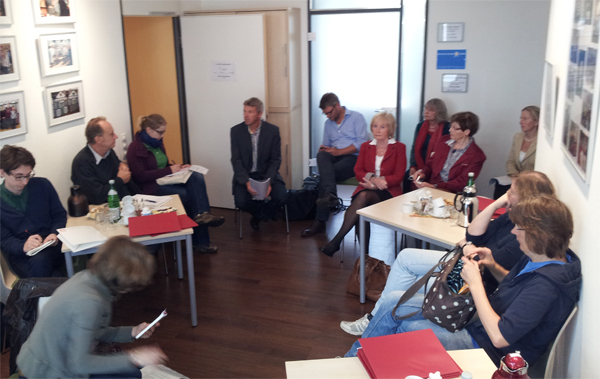 Eine Gruppe von Journalisten und Ehrenamtlichen sitzt zu einem Pressegespräch kreisförmig zusammen. Zwischen einigen Stühlen sind kleine Tische aufgestellt.