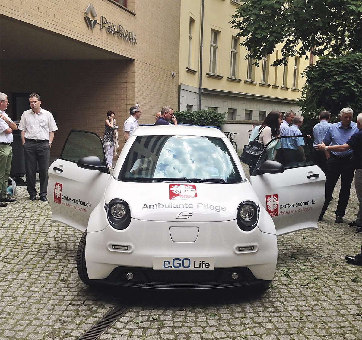 Ein weißes Elektrorauto des RCV Aachen mit geöffneten Türen, was auf einem Hinterhof inmitten einer Menschenansammlung steht