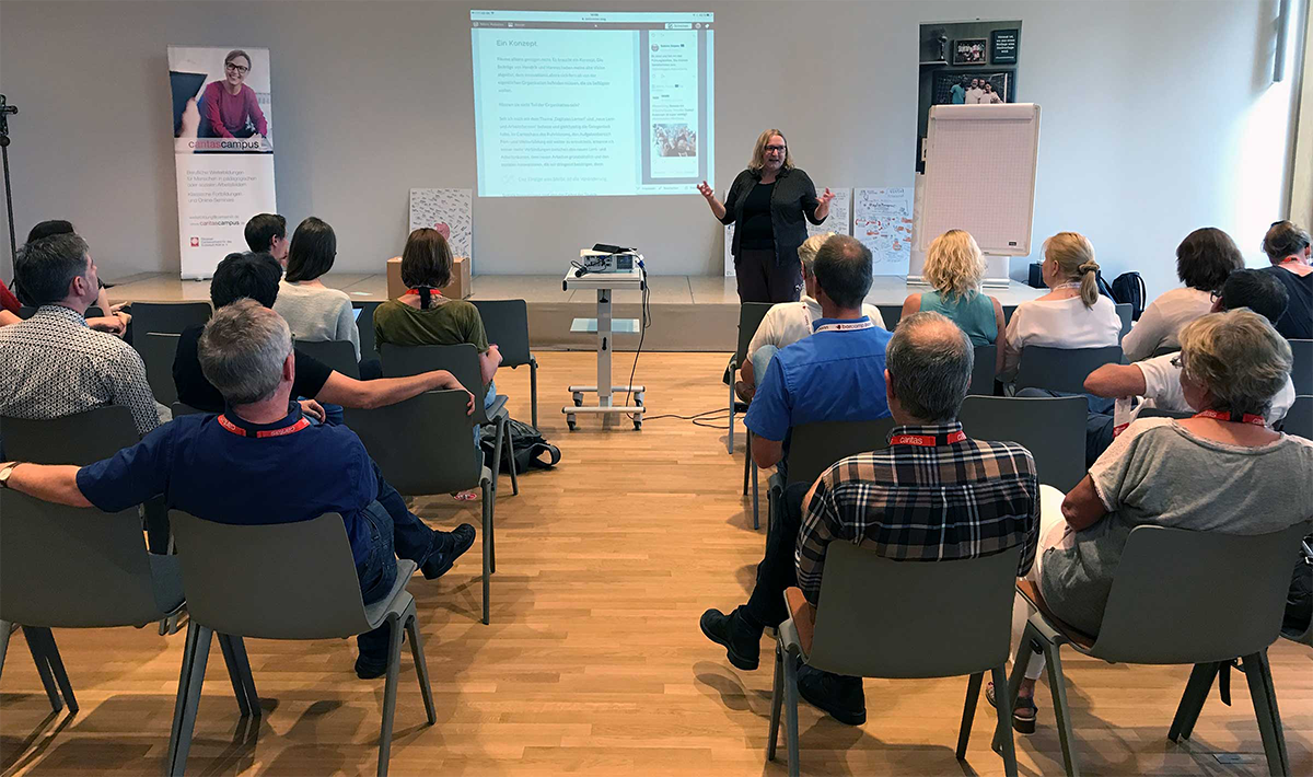 Sabine Depew steht neben einem Rollwagen, auf dem ein Beamer steht, und hält eine Präsentation auf dem Barcamp Soziale Arbeit 2017 in Siegburg. Im Vordergrund ist das Publikum zu sehen.