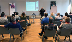 Sabine Depew bei Ihrer Präsentation Sabine Depew steht neben einem Rollwagen, auf dem ein Beamer steht, und hält eine Präsentation auf dem Barcamp Soziale Arbeit 2017 in Siegburg. Im Vordergrund ist das Publikum zu sehen.