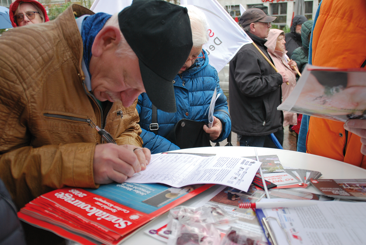 Ein Demonstrant steht an einem Stand des Aktionsbündnisses 'Wir wollen wohnen!' und unterschreibt eine Petition. Im Hintergrund sind weitere Demonstranten zu sehen.