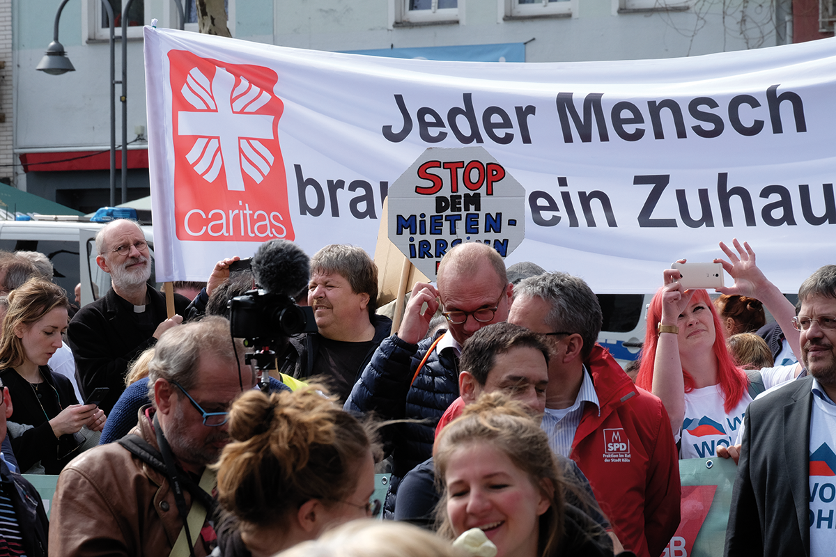 Demonstranten auf einer Demonstration an einem Aktionstag des NRW-Bündnisses 'Wir wollen wohnen!' am 6. April 2019 in Köln. Im Hintergrund ist ein großes Banner der Caritas zu sehen. 