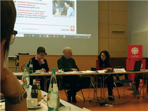 Referent/innen auf der Konferenz der Familienpflegedienste der Caritas in NRW Zwei Referentinnen und ein Referent sitzen bei der Konferenz der Familienpflegedienste der CiNW an einer Tischreihe. Im Hintergrund ist die Bühne und die Projektion eines Beamers zu sehen.