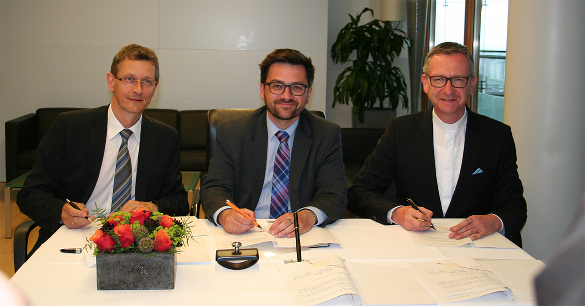 Dr. Frank Joh. Hensel, Thomas Kutschaty und Pfarrer Dr. Antonius Hamers sitzen nebeneinander an einem Tisch und unterzeichnen die Rahmenvereinbarung. Dabei blicken sie in die Kamera.