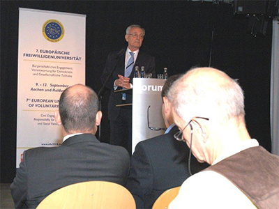 Prof. Dr. Klaus Hänsch steht an einem Rednerpult und hält eine Ansprache. Auf der Bühne ist ein Roll-up zur Europäischen Freiwilligenuniversität und im Vordergrund drei Zuschauer zu sehen.