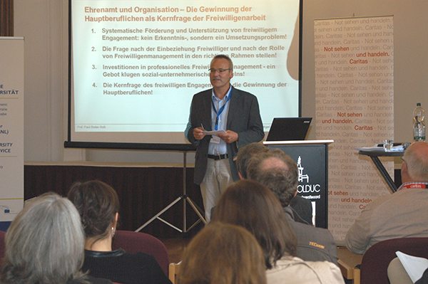 Dr. Paul-Stefan Roß steht neben einem Rednerpult und hält einen Vortrag. Hinter ihm wird seine Power-Point-Präsentation auf eine Leinwand projiziert.