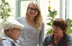 Patientenfürsprecherin mit zwei Patienten Eine Patientenfürsprecherin steht neben einem Patienten und einer Patientin ( jeweils in einer Sitzposition) und lächelt in die Kamera