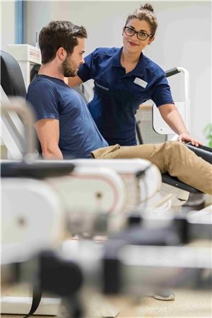 BGM Physio Eine Physiotherapeutin weist einen Patienten in die Nutzung eines Fitness-Geräts ein.